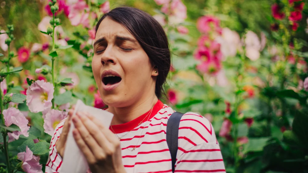 Persona en un jardín con flores de colores, llevando una camiseta de rayas rojas y blancas, sosteniendo un pañuelo cerca de la nariz y la boca.